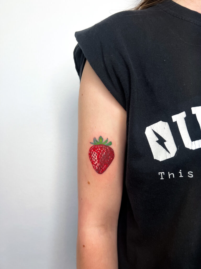 Realistic red strawberry tattoo inked on the upper arm, small and detailed color piece.