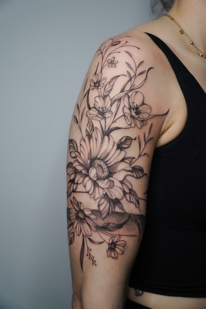 Tatuaż na ramieniu z motywem margerytek i liści, wykonany precyzyjnie w stylu fineline w studiu Fineline Tattoo Gallery.