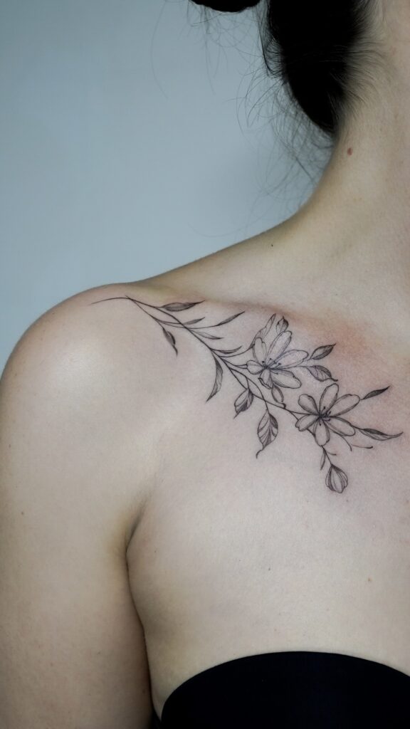 Fine line floral tattoo gracefully curving along the collarbone, showcasing delicate flowers and soft shading.