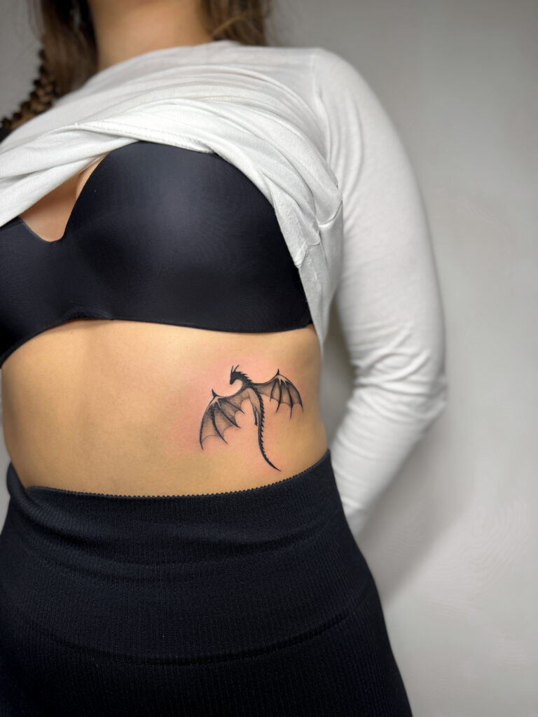 Minimalist dragon tattoo in black ink located on the side of the ribs. A simple yet bold fantasy-inspired design.
