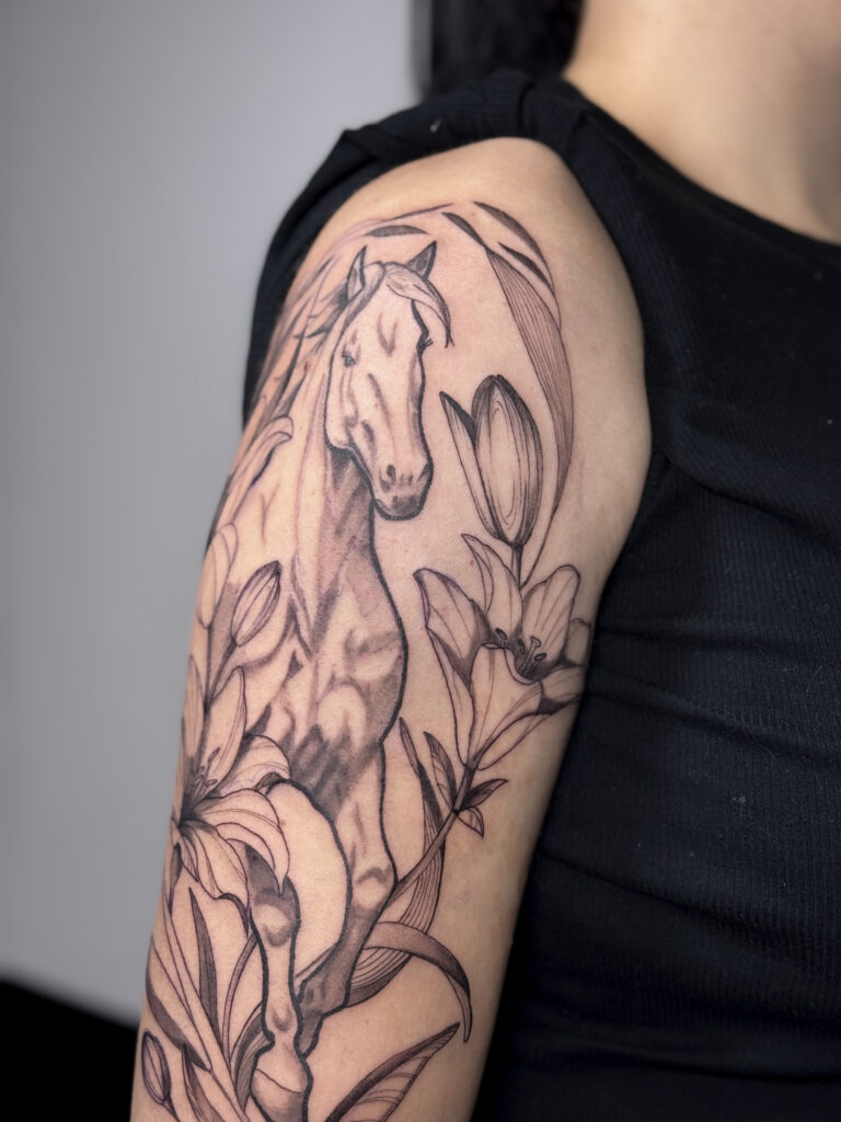Detailed upper arm tattoo of a horse surrounded by lilies and leaves in a botanical composition.