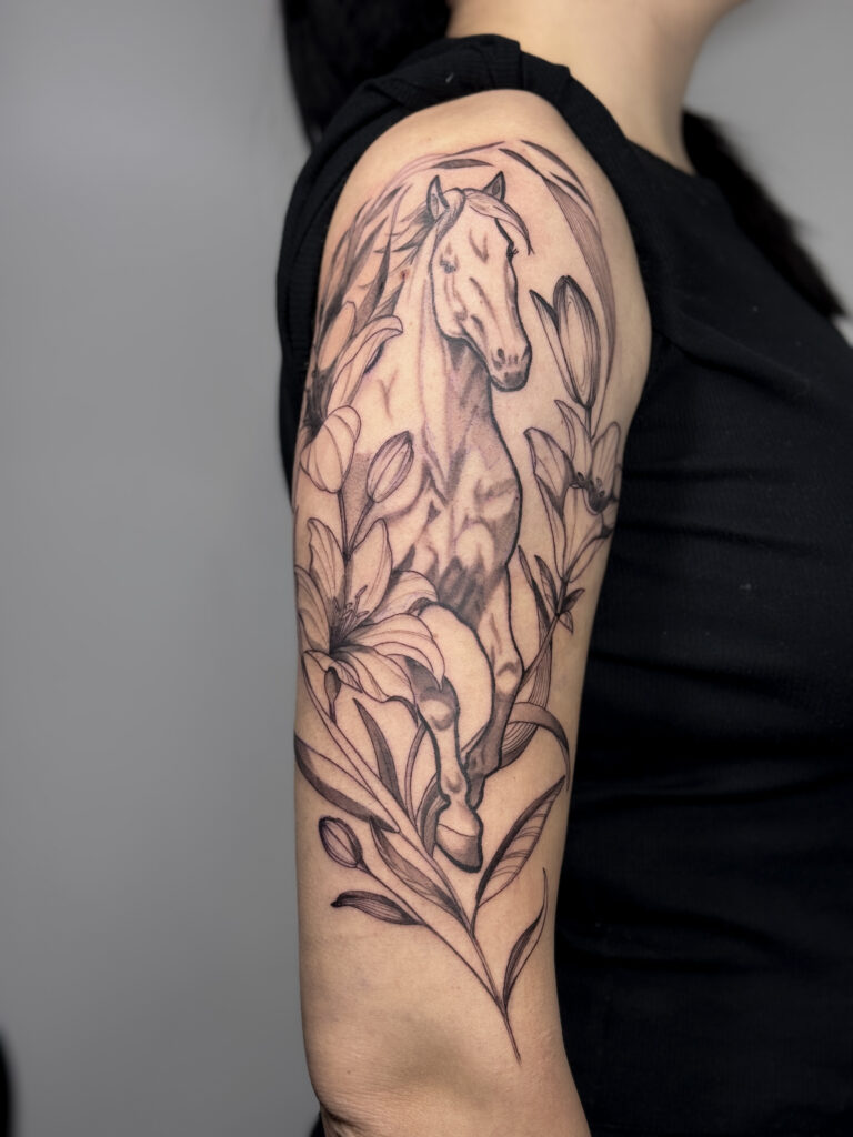 Botanical tattoo featuring a standing horse and florals wrapping the upper arm in a naturalistic layout.