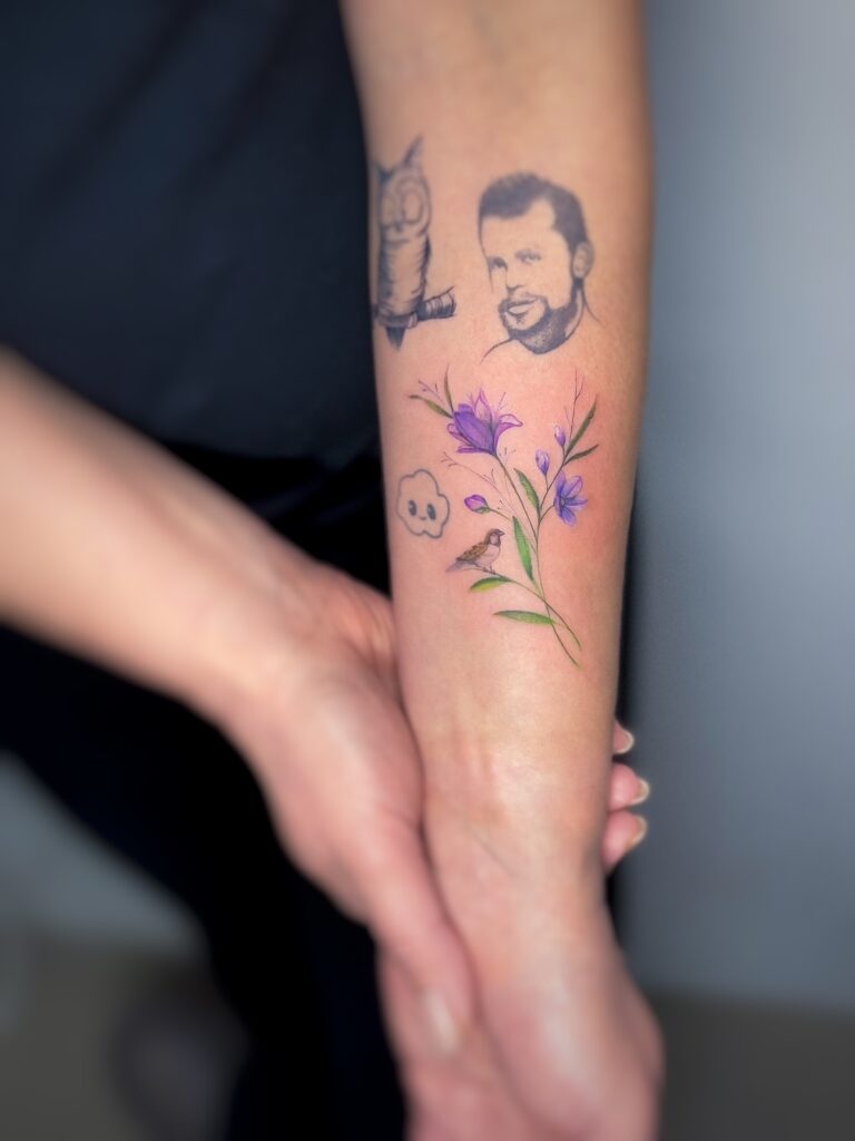 Vivid purple flower tattoo placed among existing ink on the forearm — small, vibrant, and expressive.