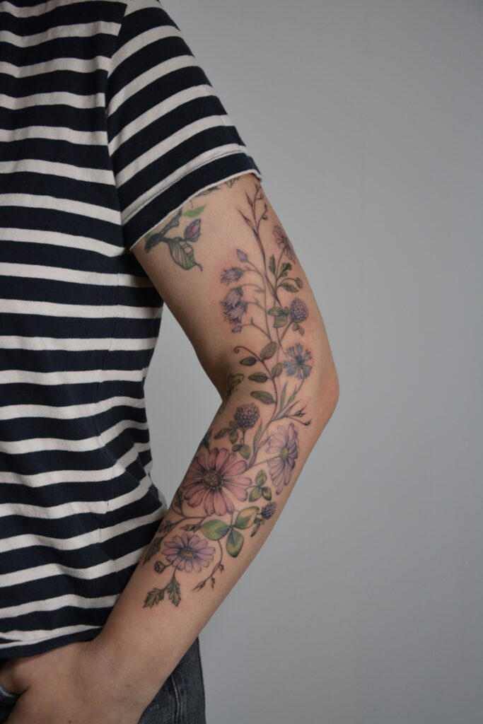 A vibrant botanical arm sleeve tattoo featuring a variety of realistic flowers in soft pastel colors. This detailed, nature-inspired design extends from the wrist to the upper arm, combining elegance with artistic expression. Perfect for lovers of floral themes and personalized body art.