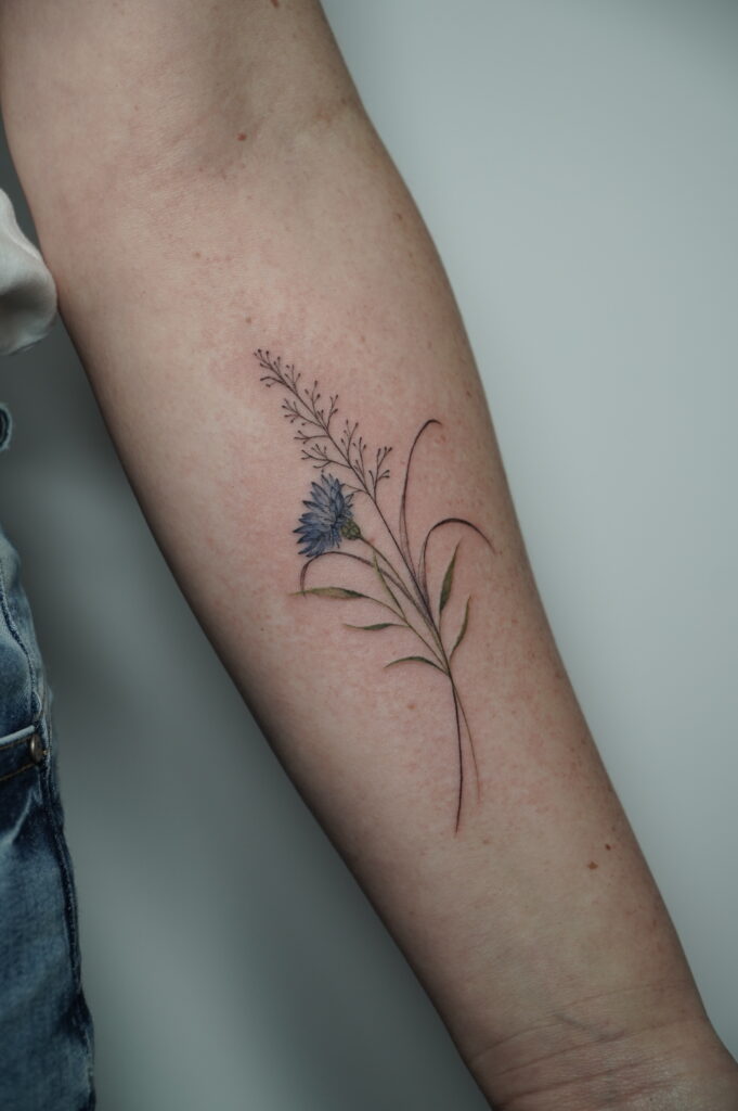 A delicate fineline forearm tattoo featuring a realistic wildflower composition with a soft blue blossom and slender botanical elements, combining elegance and nature-inspired minimalism.