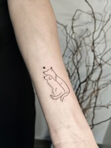 A minimalist fine line tattoo on the forearm depicting a sitting dog with a small heart above its head. A delicate and meaningful design, perfect for dog lovers or as a pet memorial tattoo. oplus_2097184