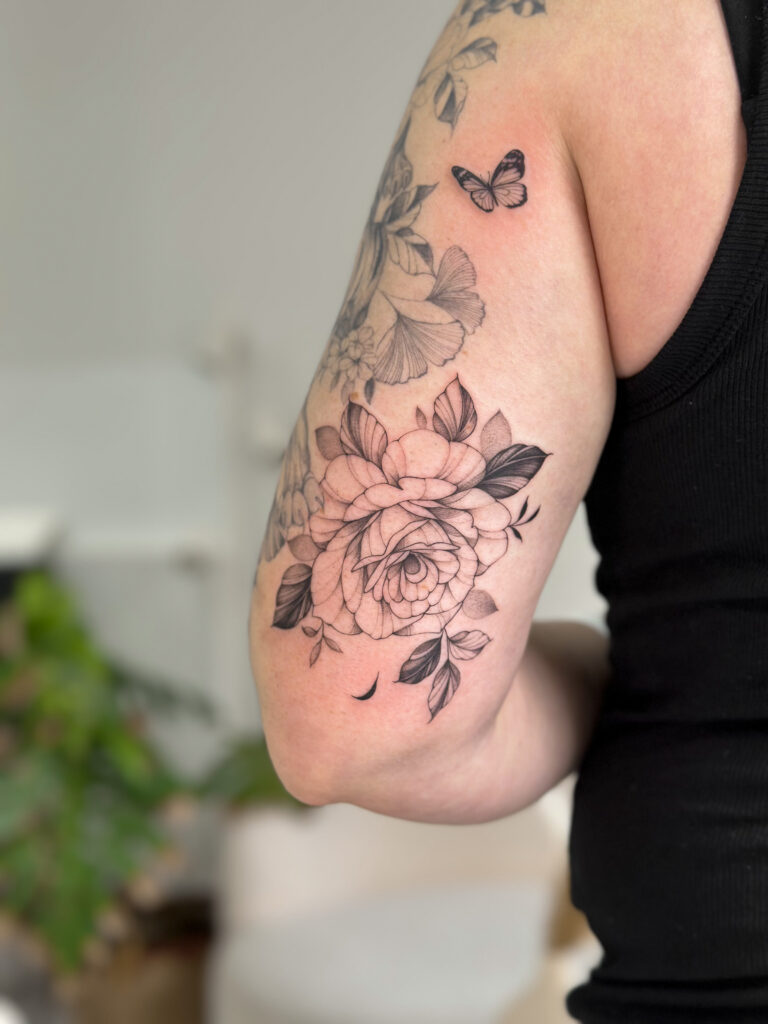 Black and grey floral tattoo featuring a detailed rose with leaves and a small butterfly, placed on the arm. A refined botanical composition with soft shading, created at Fineline Tattoo Gallery in Krakow.