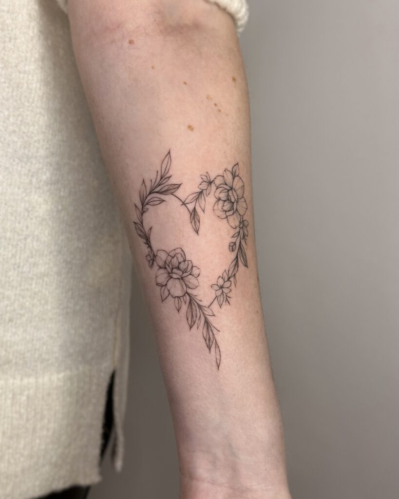 Botanical fine line floral heart tattoo in black ink placed on the forearm. A delicate and feminine composition of flowers and leaves, created at Fineline Tattoo Gallery in Krakow.
