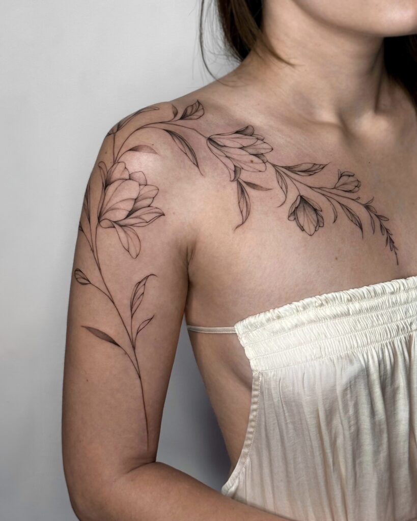 Botanical fine line floral tattoo in black ink flowing from the shoulder along the arm. A soft and feminine composition of blooming flowers and leaves, created at Fineline Tattoo Gallery in Krakow.