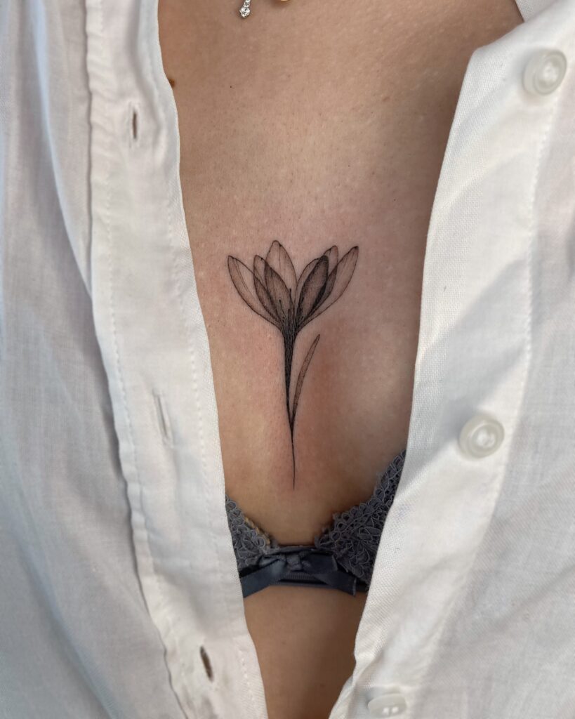 Botanical fine line floral tattoo in black ink placed between the breasts along the sternum. A delicate and elegant minimal flower design created at Fineline Tattoo Gallery in Krakow.