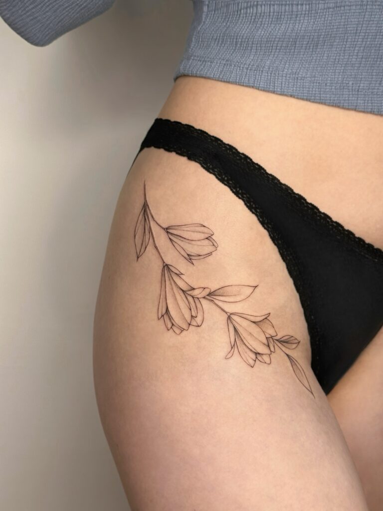 Botanical fine line floral tattoo in black ink placed on the thigh. A delicate and feminine composition of flowing flowers and leaves, created at Fineline Tattoo Gallery in Krakow.