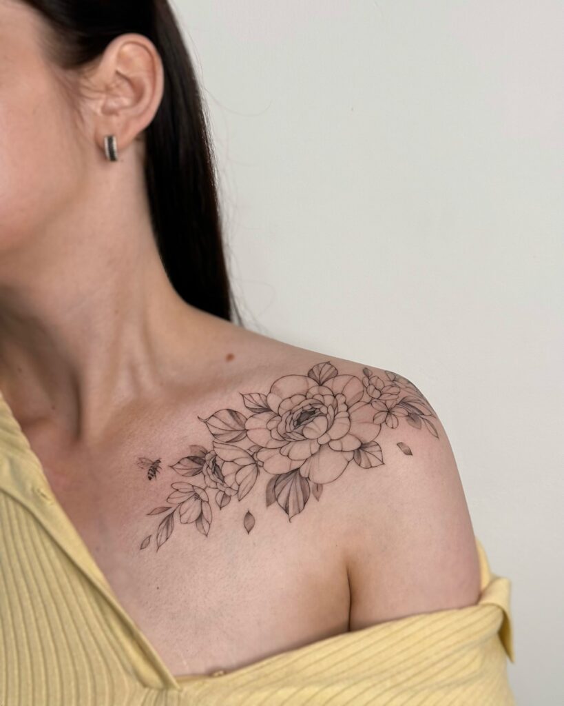 Botanical fine line peony tattoo with delicate leaves and a small bee, placed across the shoulder and collarbone. A soft and feminine black ink floral composition created at Fineline Tattoo Gallery in Krakow.