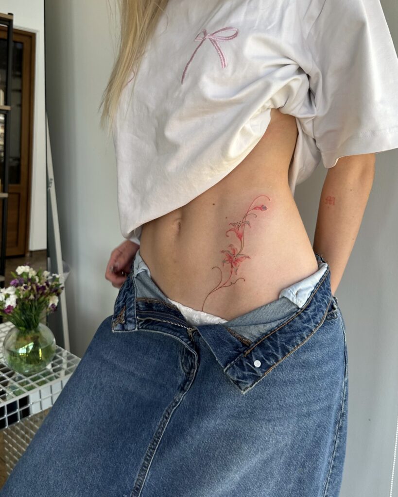 Delicate fine line red floral tattoo placed on the side of the body. Elegant and feminine design created at Fineline Tattoo Gallery in Krakow.