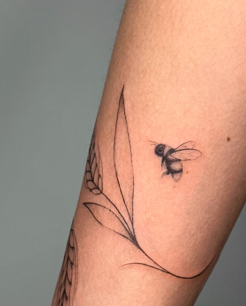 A small fine line bee tattoo paired with soft botanical linework. The bee is lightly shaded, giving it a gentle and natural look, while the surrounding leaves add flow and elegance. A subtle, nature-inspired piece that feels airy and graceful.