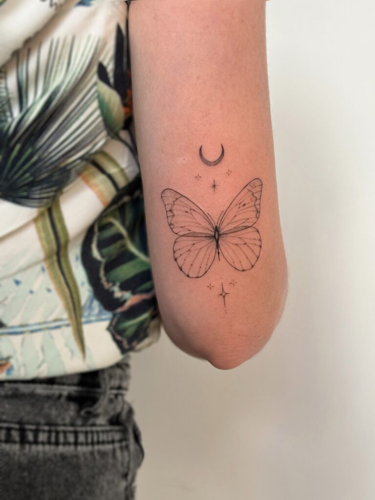 Fine line butterfly tattoo with a crescent moon and small star accents, placed on the arm. A delicate and symbolic black ink design created at Fineline Tattoo Gallery in Krakow.