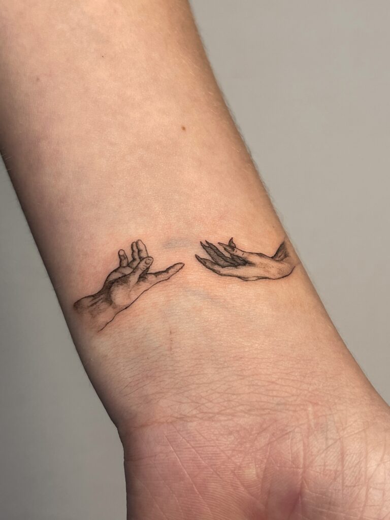 Fine line tattoo of two hands reaching toward each other, placed on the wrist. A delicate and symbolic black ink design representing connection and closeness, created at Fineline Tattoo Gallery in Krakow.