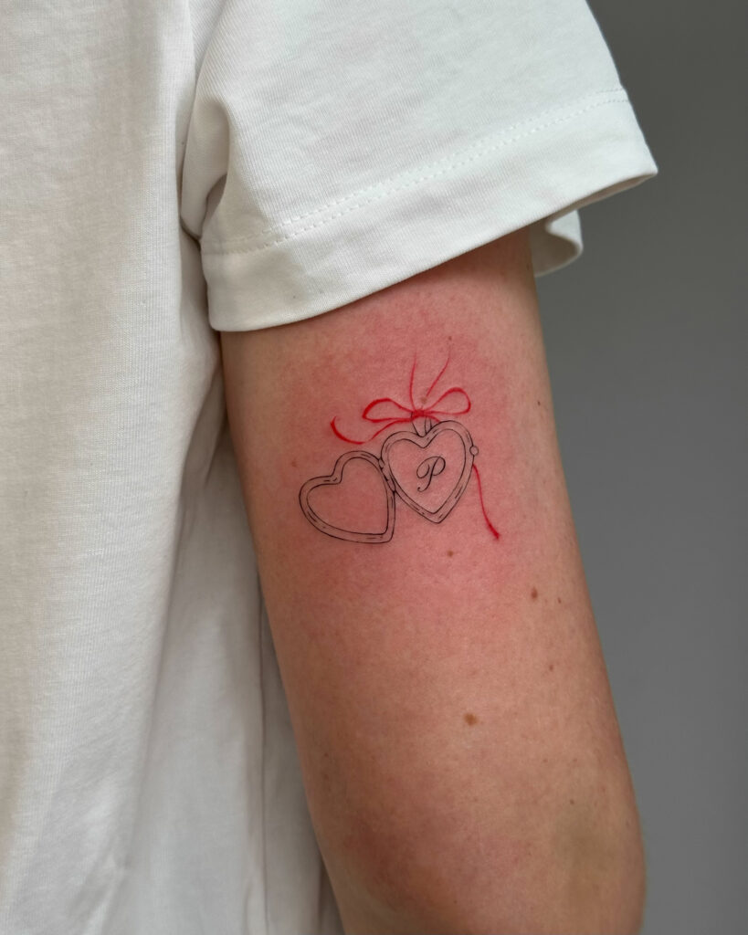 Romantic fine line heart locket tattoo with a red bow, placed on the arm. A delicate and symbolic black and red ink design created at Fineline Tattoo Gallery in Krakow.
