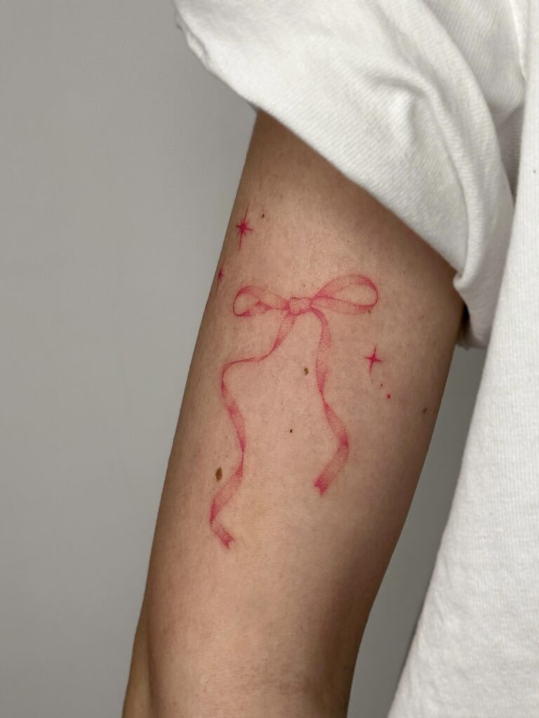 Fine line red bow tattoo placed on the arm, featuring soft ribbon details and subtle star accents. A delicate and feminine red ink design created at Fineline Tattoo Gallery in Krakow