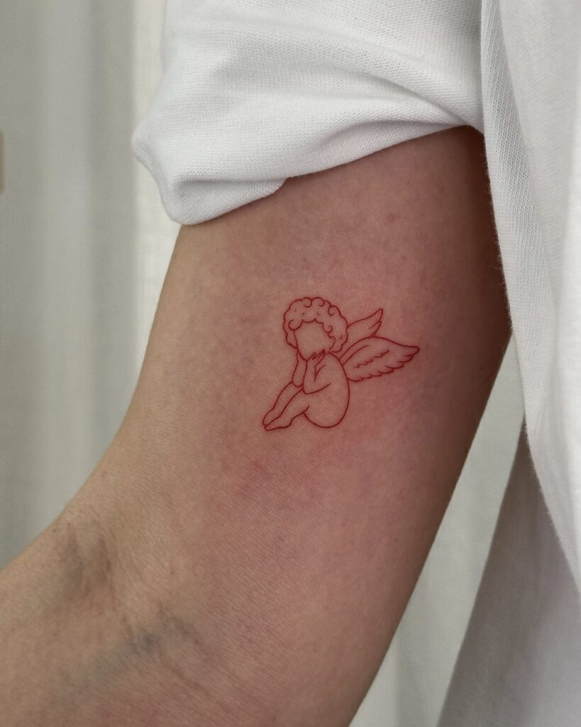 Fine line red ink cherub tattoo placed on the arm. A delicate, soft and symbolic design created at Fineline Tattoo Gallery in Krakow.