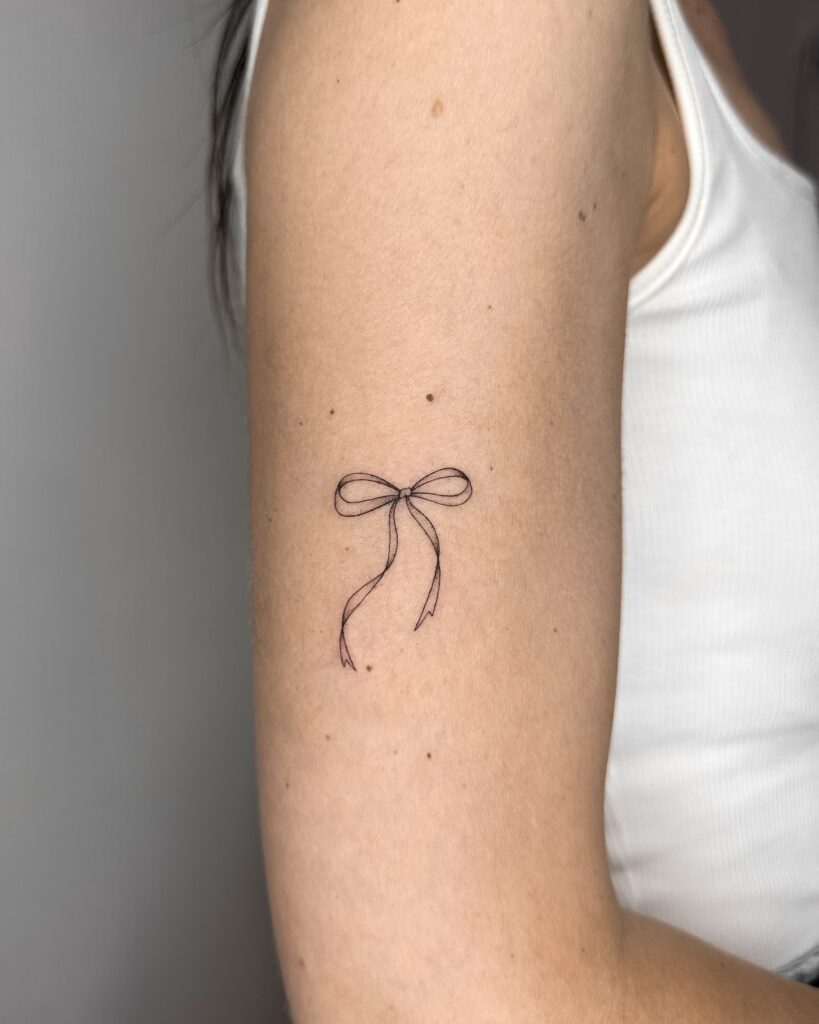 Minimal fine line ribbon bow tattoo in black ink placed on the arm. A delicate, soft and feminine design created at Fineline Tattoo Gallery in Krakow.