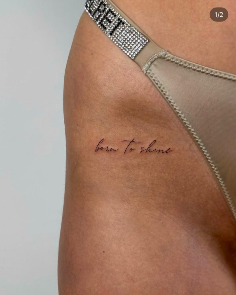 Fine line script tattoo reading “born to shine” placed on the hip. A minimal and elegant lettering design in soft red ink, created at Fineline Tattoo Gallery in Krakow.