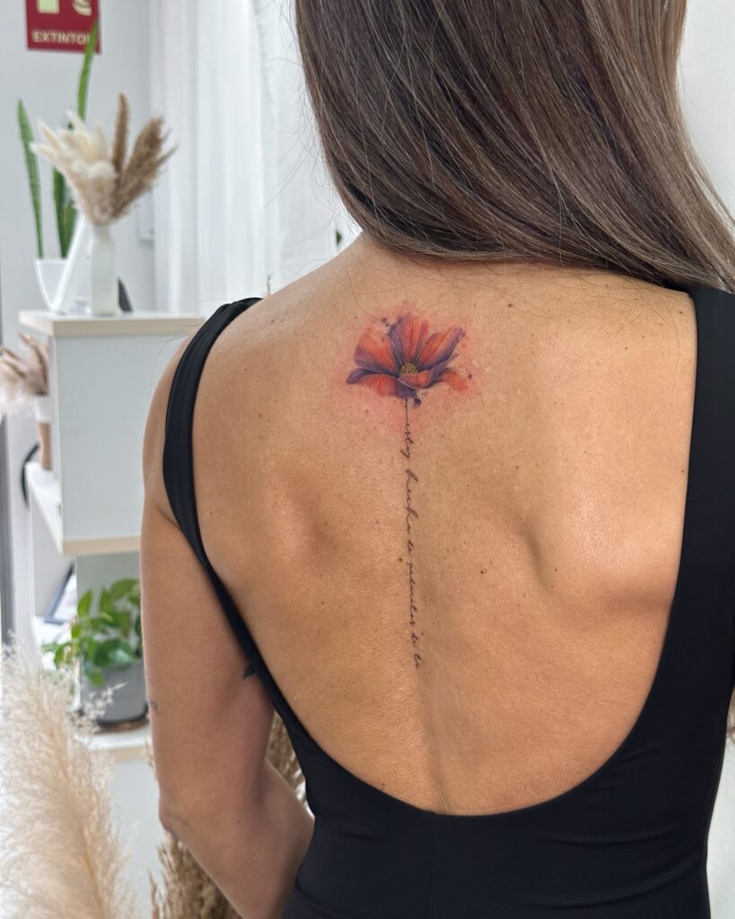 Fine line floral tattoo with soft watercolor shading placed along the upper spine. A delicate and feminine flower design with subtle lettering, created at Fineline Tattoo Gallery in Krakow.