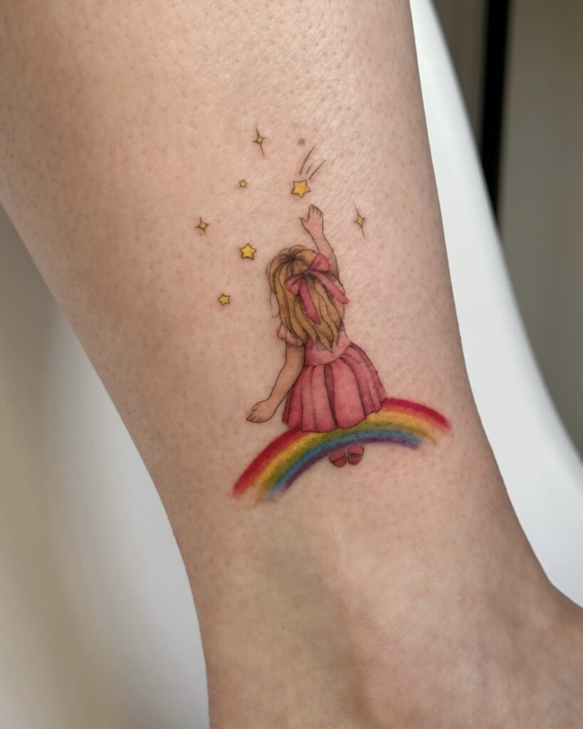 Colorful tattoo of a little girl in a pink dress reaching for stars while standing on a rainbow. A soft, dreamy and joyful design created at Fineline Tattoo Gallery in Krakow.