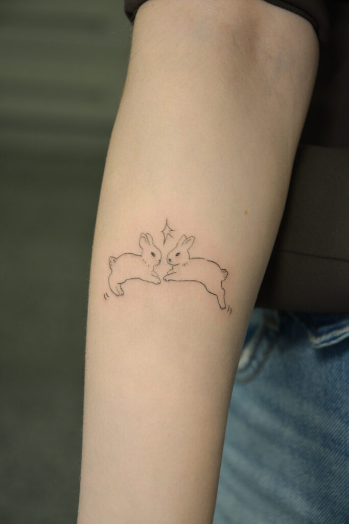 Subtelny tatuaż dwóch królików w stylu fine line wykonany przez kursantkę podczas szkolenia Mini Tattoo w Fineline Tattoo Gallery. Idealny przykład nauki delikatnych i minimalistycznych tatuaży.