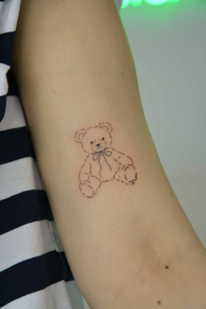 Delikatny mini tatuaż misia wykonany podczas szkolenia Mini Tattoo w Fineline Tattoo Gallery. Przykład pracy kursantki w stylu fine line, idealny jako mały i subtelny projekt dla początkujących tatuatorów.