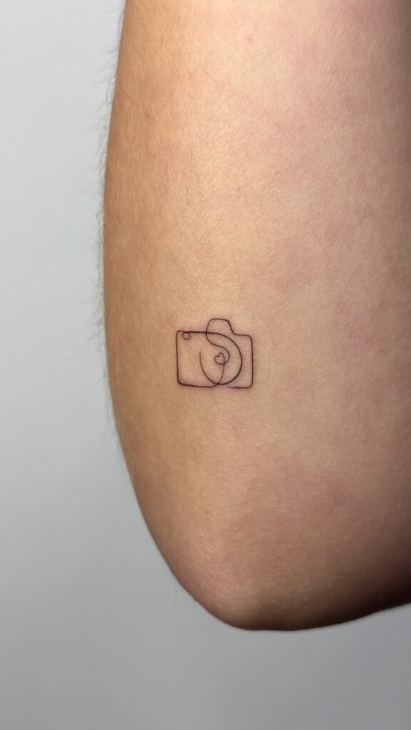 Minimalist fine line camera tattoo with a small heart detail, placed on the arm. A subtle and personal design created at Fineline Tattoo Gallery in Krakow.