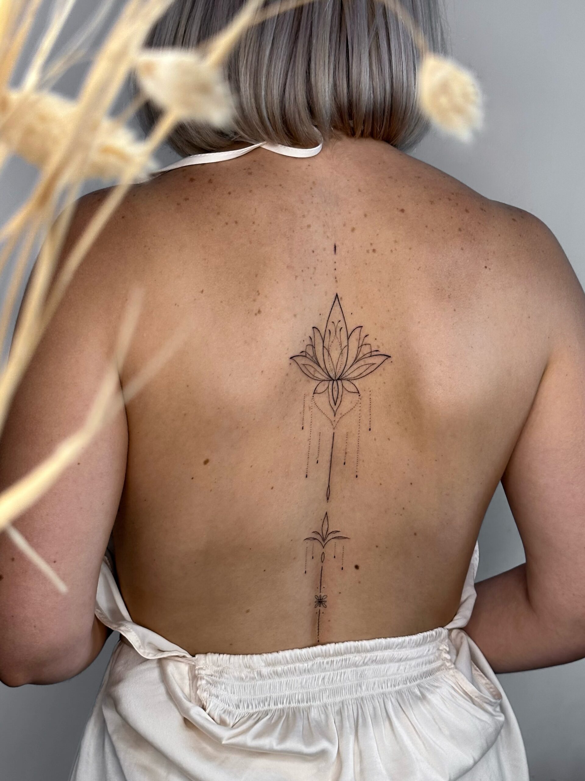 Ornamental fine line lotus tattoo running along the spine. A delicate and feminine black ink design with botanical and geometric elements, created at Fineline Tattoo Gallery in Krakow.