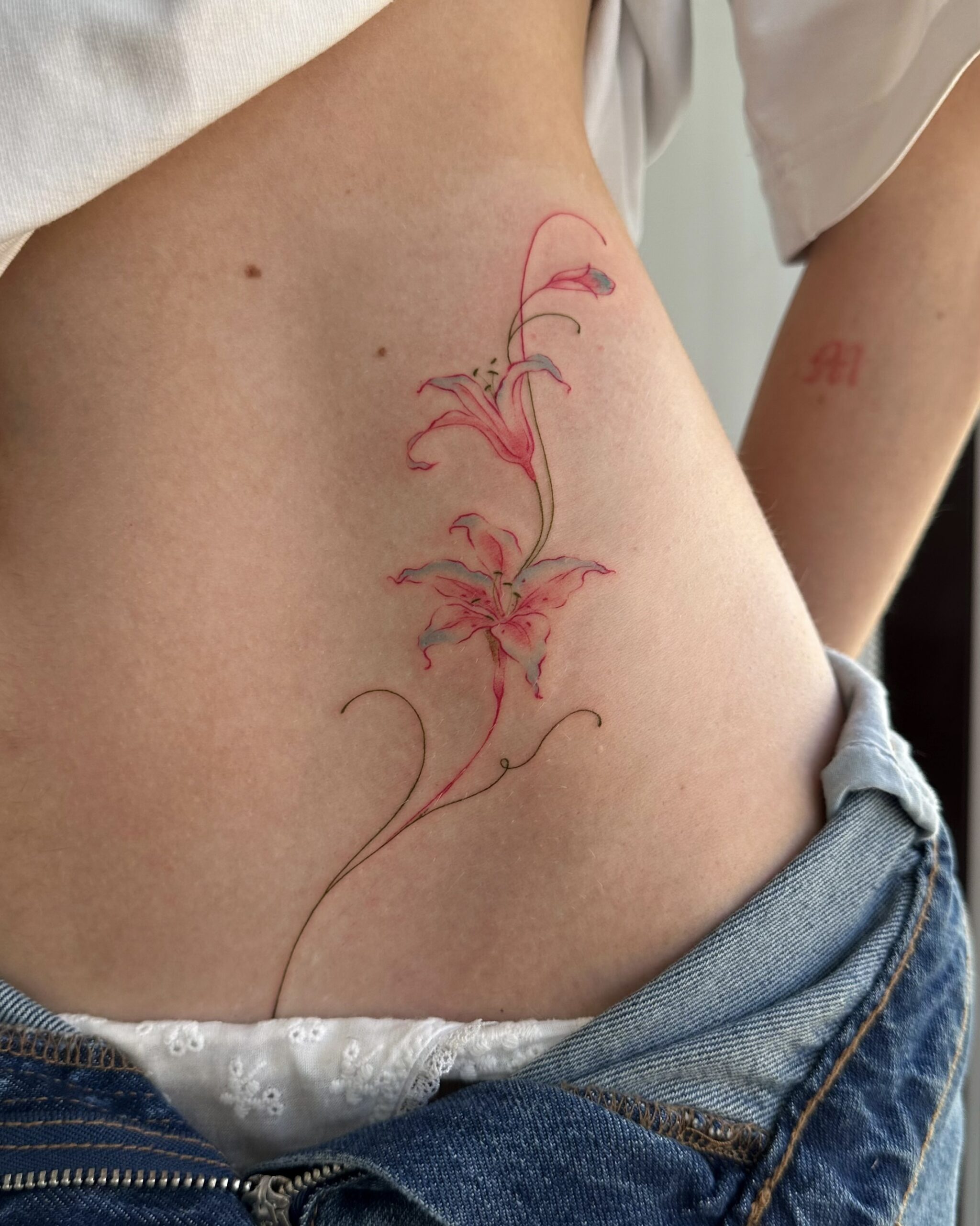 Close-up of a delicate red lily tattoo in fine line style, placed on the side of the body. A feminine and elegant floral design created at Fineline Tattoo Gallery in Krakow.