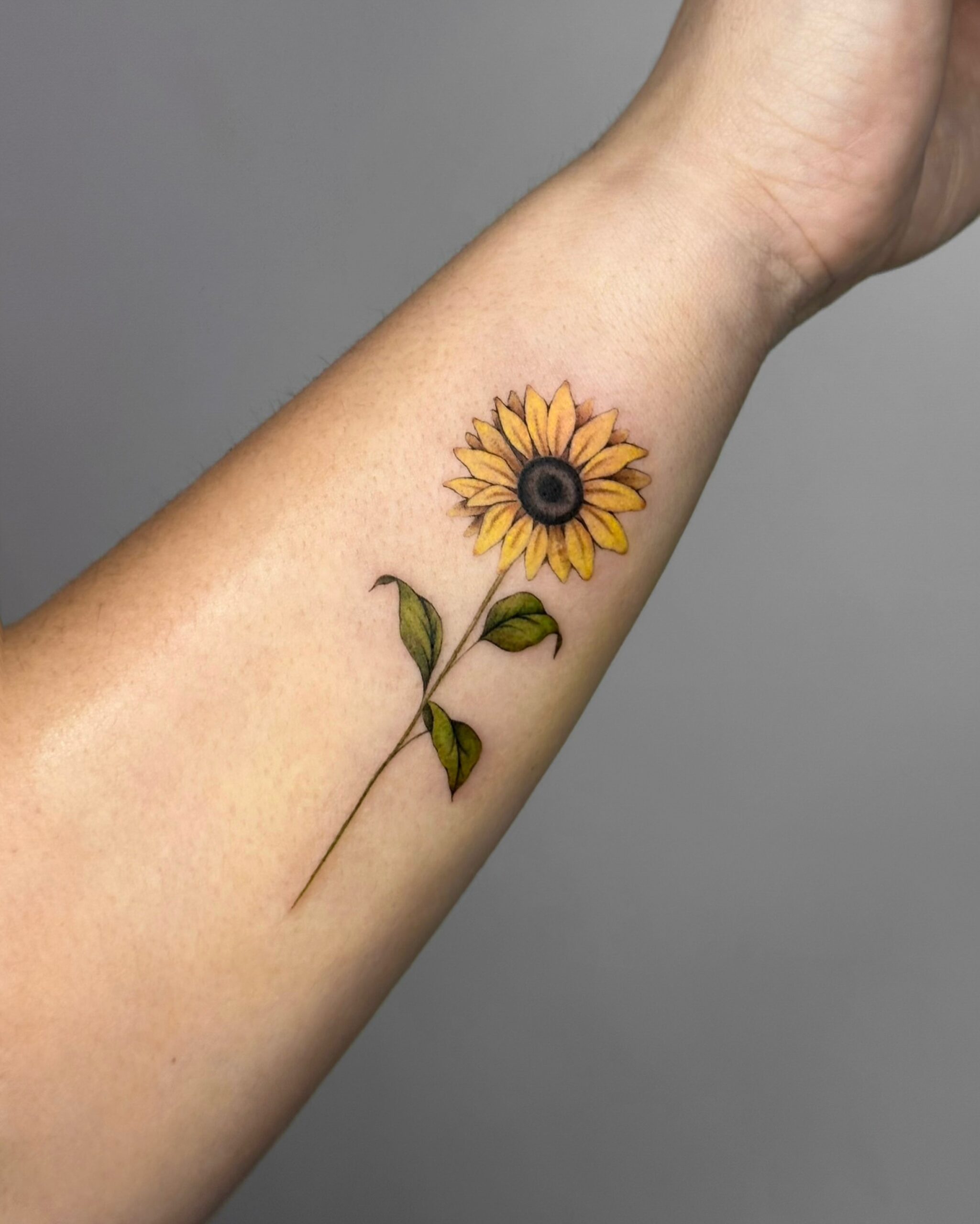 A delicate fine line sunflower tattoo with soft color shading. The warm yellows and greens create a gentle, natural look, while the slim stem and subtle details keep the design light and elegant. A bright, uplifting piece symbolizing warmth, positivity and light.