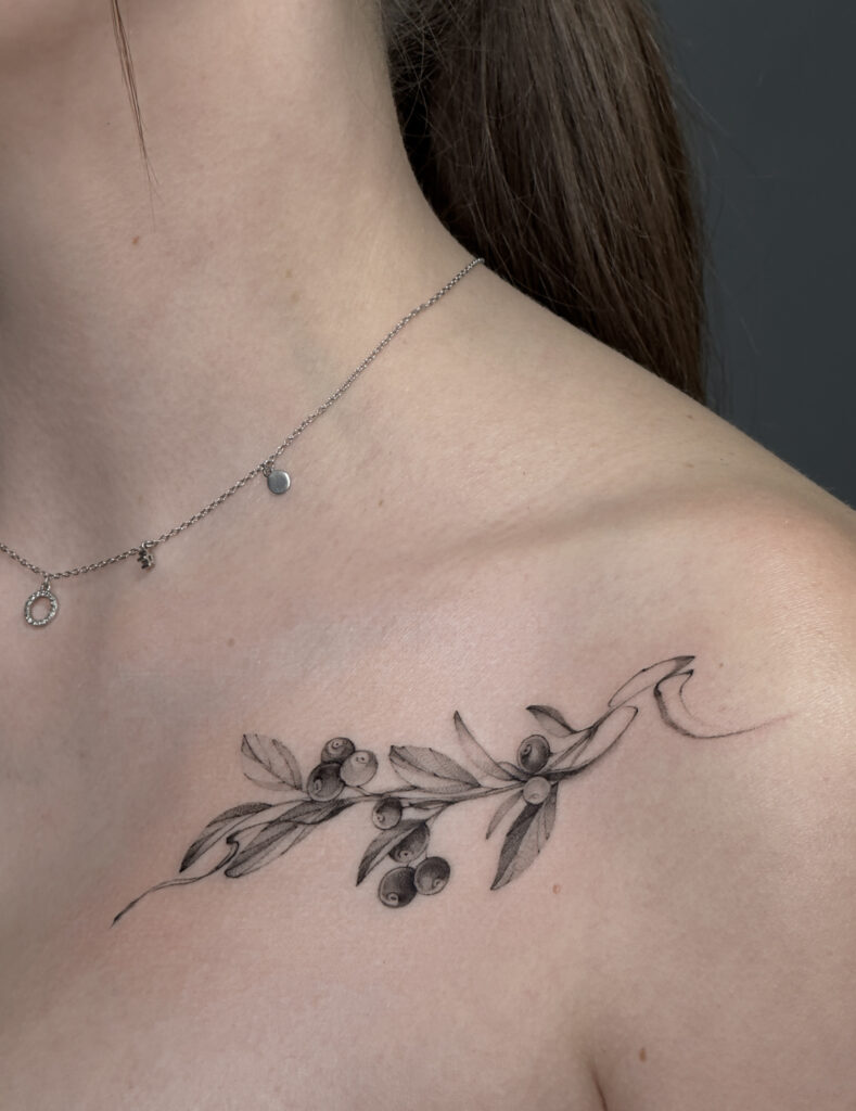Delikatny tatuaż fine line przedstawiający gałązkę oliwną z liśćmi i owocami, wykonany cienką linią na obojczyku. Subtelny, minimalistyczny tatuaż botaniczny w czerni, idealny dla osób ceniących eleganckie i kobiece wzory.