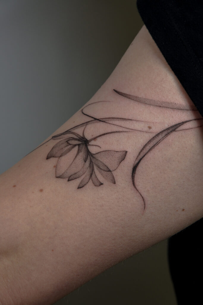 Delikatny tatuaż fine line z motywem pojedynczego kwiatu, wykonany cienką linią i subtelnym cieniowaniem typu dotwork na ramieniu. Minimalistyczny, botaniczny tatuaż w czerni, podkreślający lekkość i elegancję formy.