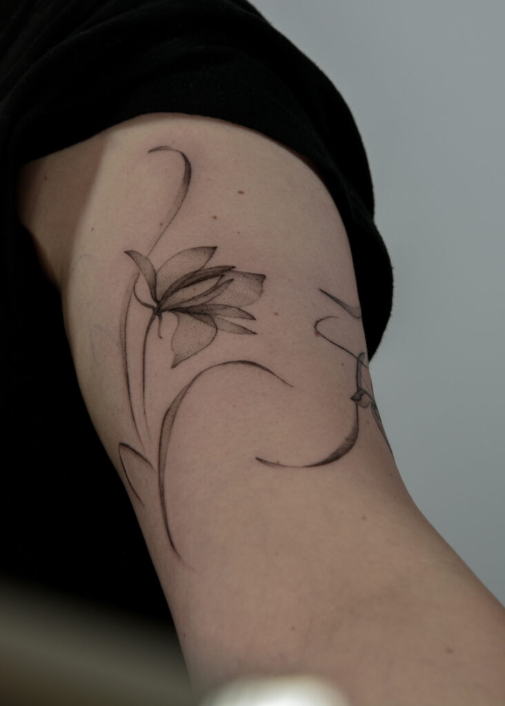 Delikatny tatuaż fine line z motywem kwiatowym, wykonany cienką linią i subtelnym cieniowaniem typu dotwork na ramieniu. Minimalistyczny, kobiecy tatuaż botaniczny w czerni, podkreślający naturalne linie ciała.