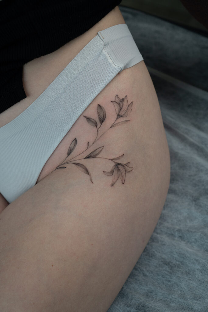 Delikatny tatuaż kwiatowy w stylu fine line z motywem gałązki i kwiatów, wykonany cienką linią z subtelnym cieniowaniem na udzie. Minimalistyczny, botaniczny tatuaż w czerni, elegancko podkreślający linię ciała.