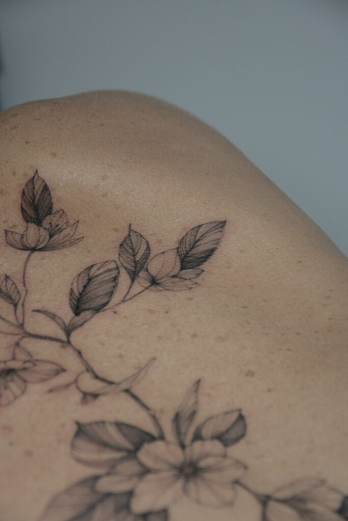 Delikatny tatuaż botaniczny w stylu fine line z motywem liści i kwiatów, wykonany cienką linią z subtelnym cieniowaniem na ramieniu. Elegancki, minimalistyczny tatuaż w czerni, harmonijnie dopasowany do naturalnych kształtów ciała.