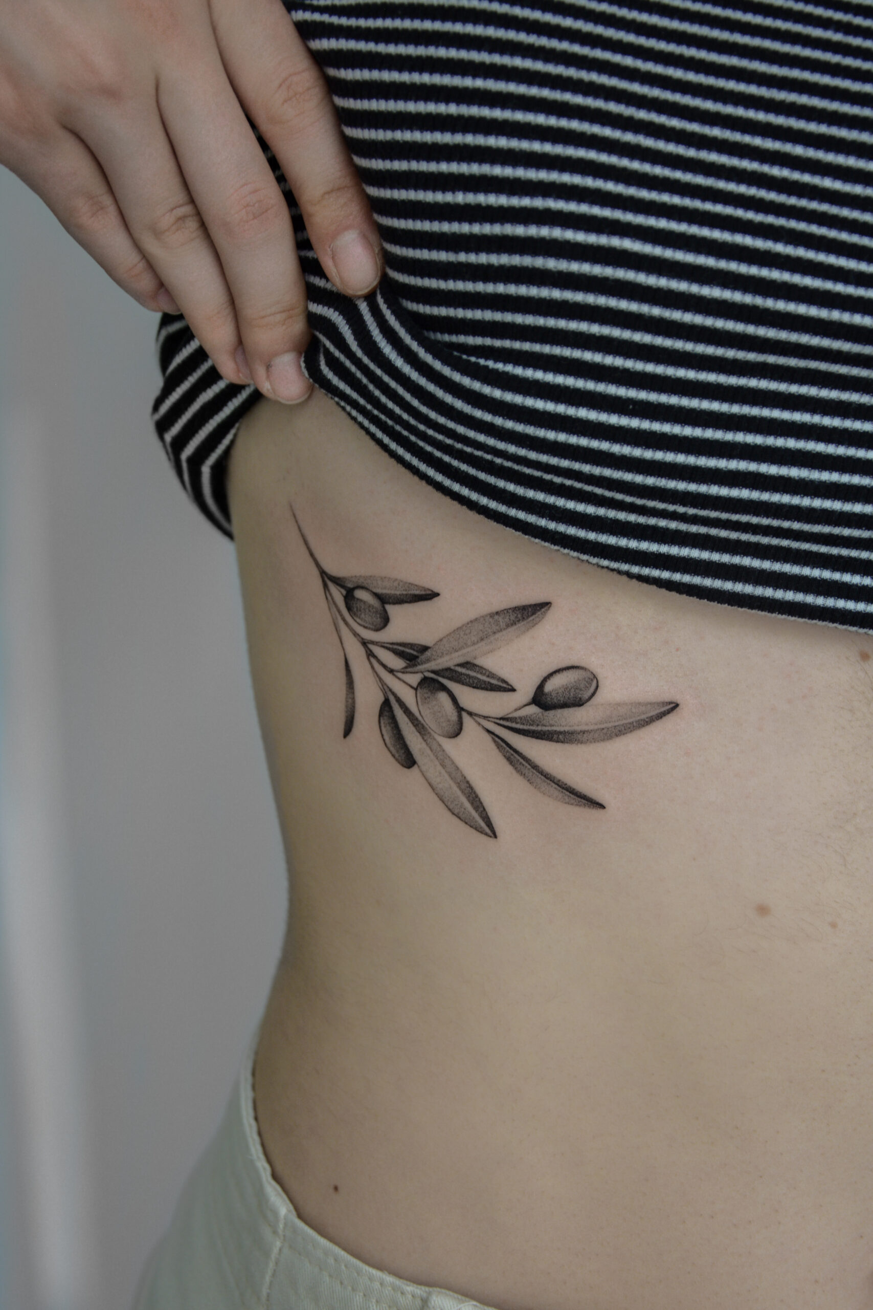Delikatny tatuaż fine line z motywem gałązki oliwnej i oliwek, wykonany cienką linią z subtelnym cieniowaniem typu dotwork na boku ciała. Minimalistyczny, botaniczny tatuaż w czerni, dyskretnie podkreślający linię sylwetki.