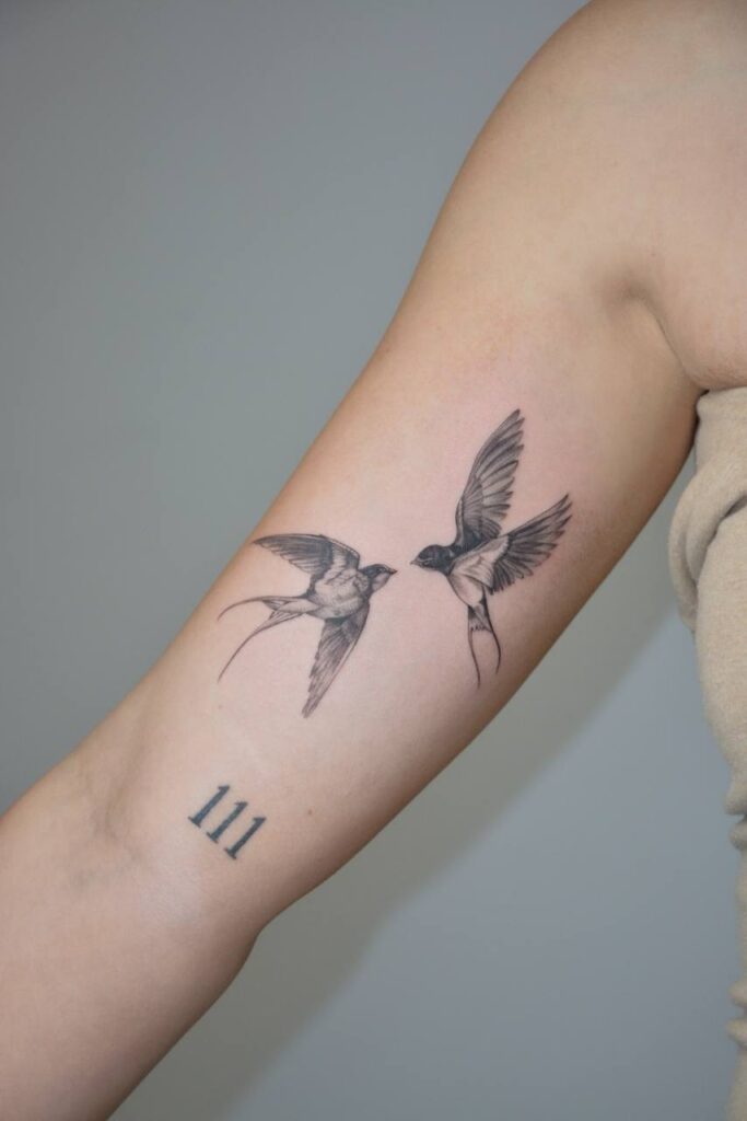 Delikatny tatuaż przedstawiający dwie jaskółki w locie, wykonany w stylu fine line na przedramieniu. Symbol wolności, miłości i powrotu do domu – subtelna forma z precyzyjnym cieniowaniem.