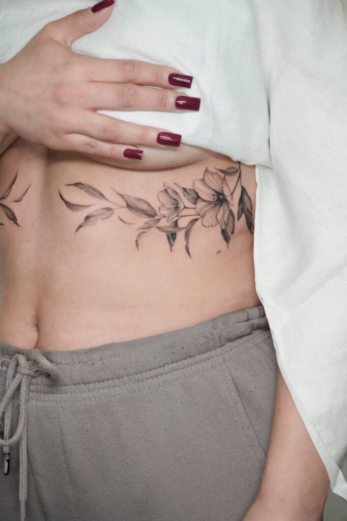 Czarno-szary tatuaż kwiatowy w stylu fine line umieszczony na boku tułowia. Subtelny, kobiecy motyw z delikatnymi liśćmi i kwiatami, idealny dla miłośniczek eleganckich, minimalistycznych kompozycji.
