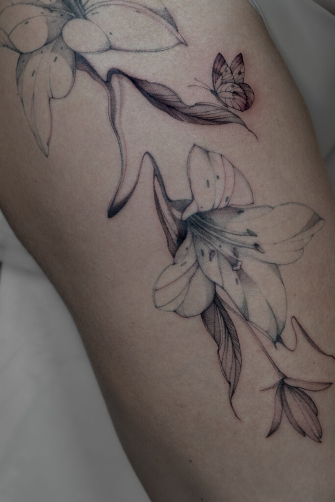 Delikatny tatuaż fine line z motywem lilii i małego motyla, wykonany cienką linią z subtelnym cieniowaniem typu dotwork. Elegancki tatuaż botaniczny w czerni, podkreślający lekkość kompozycji i naturalny ruch form.