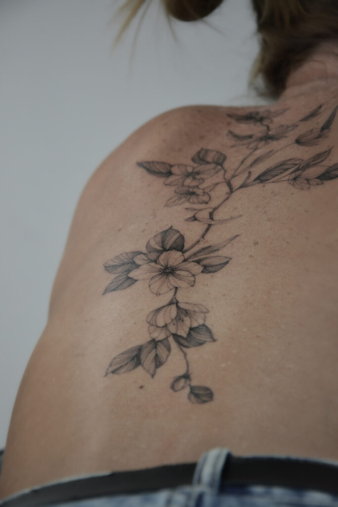 Delikatny tatuaż kwiatowy w stylu fine line z motywem gałązki kwiatowej, wykonany cienką linią i subtelnym cieniowaniem na plecach. Elegancki tatuaż botaniczny w czerni, harmonijnie dopasowany do naturalnych linii ciała.