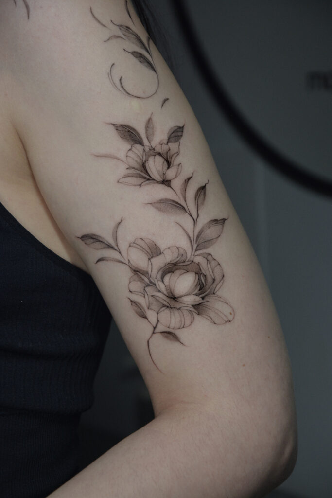 Delikatny tatuaż kwiatowy w stylu fine line z motywem kwiatów i liści, wykonany cienką linią z subtelnym cieniowaniem na ramieniu. Elegancki, botaniczny tatuaż w czerni, harmonijnie dopasowany do kształtu ręki.