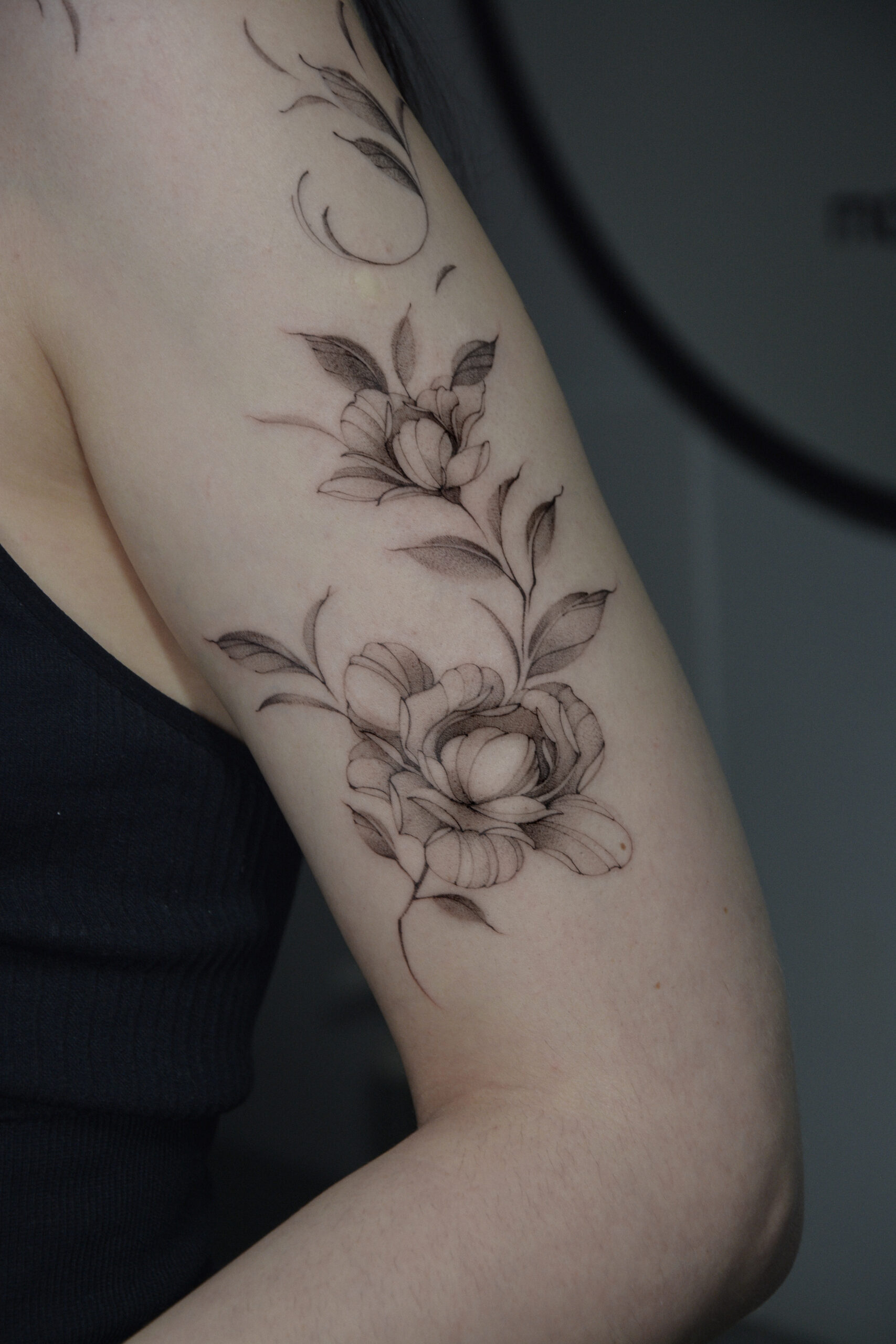 Delikatny tatuaż kwiatowy w stylu fine line z motywem kwiatów i liści, wykonany cienką linią z subtelnym cieniowaniem na ramieniu. Elegancki, botaniczny tatuaż w czerni, harmonijnie dopasowany do kształtu ręki.