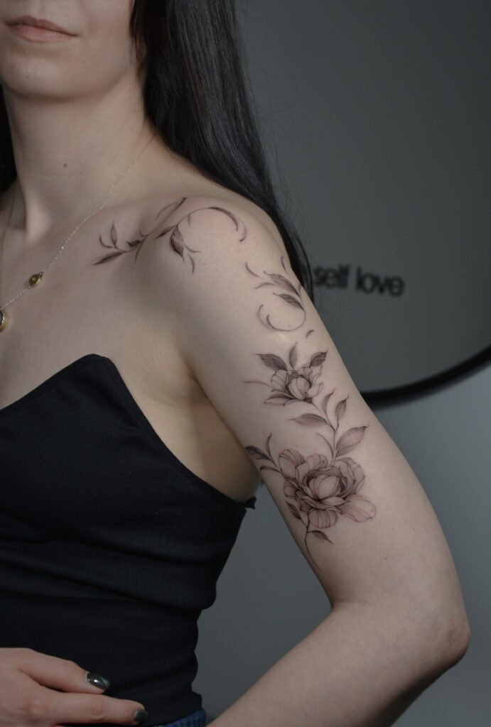 Elegancki tatuaż kwiatowy w stylu fine line, wykonany cienką linią z delikatnym cieniowaniem, obejmujący ramię i górną część ręki. Subtelny tatuaż botaniczny w czerni, podkreślający kobiecą formę i naturalny ruch linii.