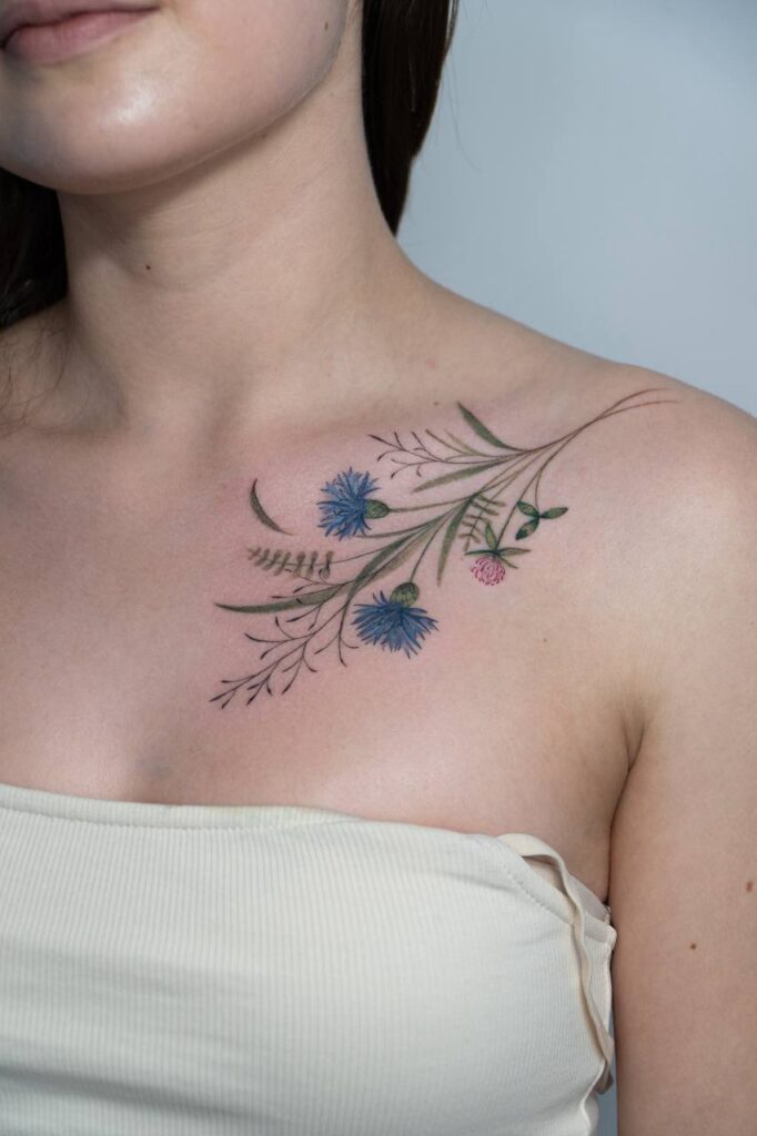 Delikatny, kolorowy tatuaż kwiatowy na obojczyku — finezyjne chabry i subtelne botaniczne gałązki w stylu fine line. Idealna propozycja dla osób szukających kobiecego, naturalnego wzoru.