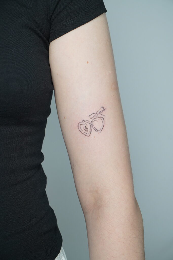 Delikatny tatuaż w stylu fine line przedstawiający dwa serduszka zawieszone na wstążce. Minimalistyczny wzór wykonany na przedramieniu – idealny wybór dla osób szukających symbolicznego i subtelnego tatuażu.
