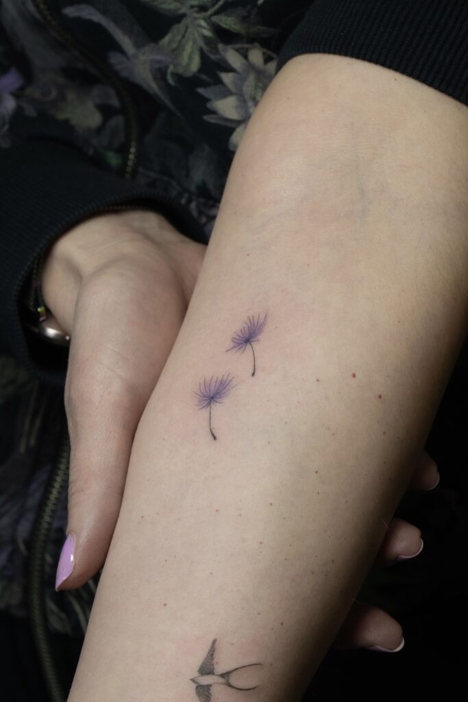 Niezwykle delikatny i minimalistyczny tatuaż przedstawiający nasiona dmuchawca w subtelnym fioletowym odcieniu. Praca wykonana w technice fine line przez Olgę (@olga_sankova_tattoo) w krakowskim studiu Fineline Tattoo Gallery. Tatuaż na przedramieniu charakteryzuje się lekkością i precyzją, będąc subtelną ozdobą kobiecego ciała.