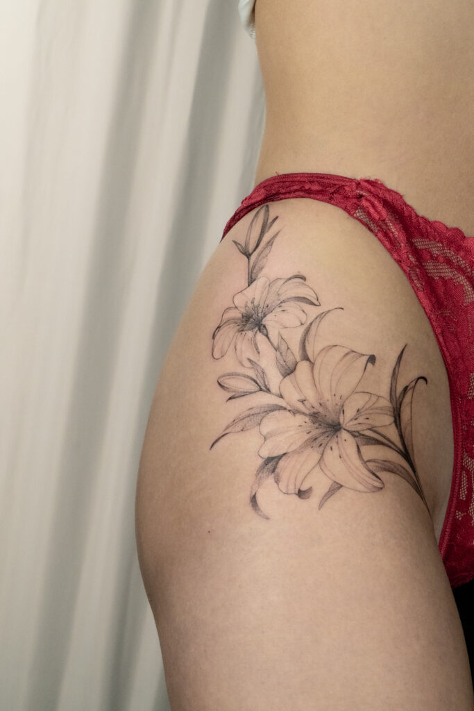 Delikatny, wielkoformatowy tatuaż kwiatowy na biodrze przedstawiający lilie. Praca wykonana przez Olgę (@olga_sankova_tattoo) w krakowskim studiu Fineline Tattoo Gallery. Kompozycja wykonana w technice cienkiej linii z miękkim cieniowaniem, zaprojektowana tak, aby podkreślać naturalne linie kobiecej sylwetki.